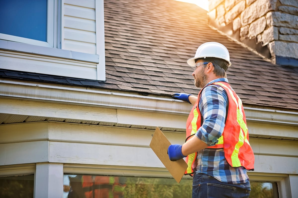 Roofing surveyor in a hard hat inspecting the roof of a house from a ladder
